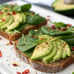 A close-up image of Cleansing Avocado & Spinach Toast with creamy avocado slices and fresh spinach on whole-grain bread, garnished with chili flakes.